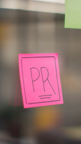 Vertical-Video-Close-Up-Of-Woman-Putting-Stick-Note-With-PR-On-It-Onto-To-Transparent-Screen-In-Office